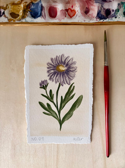 A hand-painted botanical art print of a purple aster flower with green leaves on textured white paper, displayed next to a red-handled paintbrush on a light wood surface with a watercolor palette visible at the top.