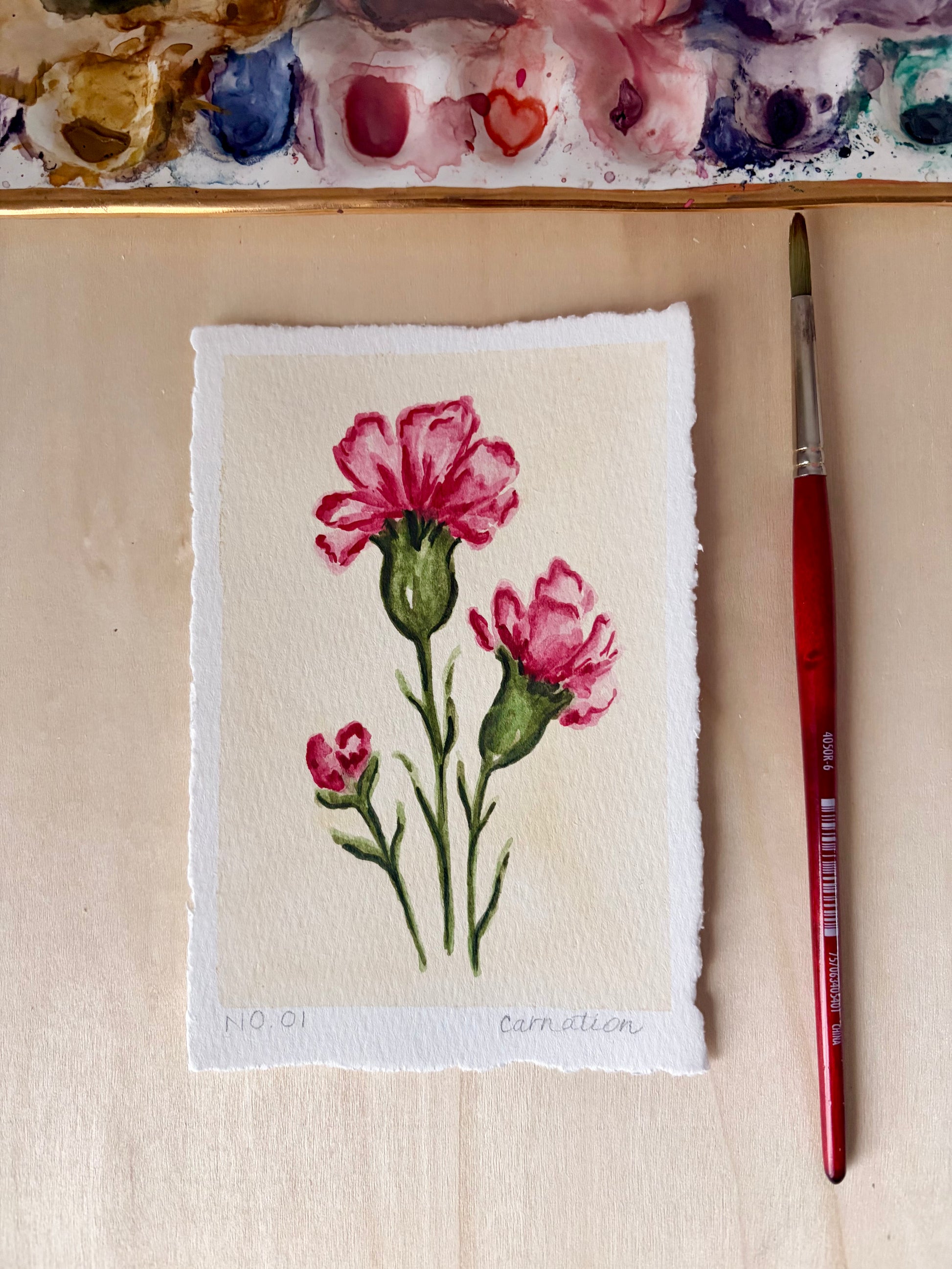 A small handmade paper painting of two pink carnations with green stems, displayed on a light wood surface next to a red-handled paintbrush and a watercolor palette.