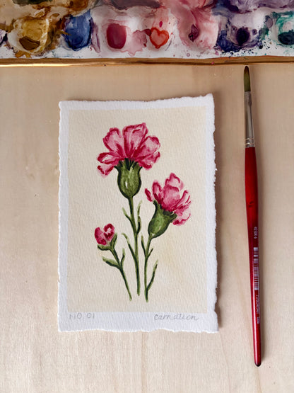 A small handmade paper painting of two pink carnations with green stems, displayed on a light wood surface next to a red-handled paintbrush and a watercolor palette.