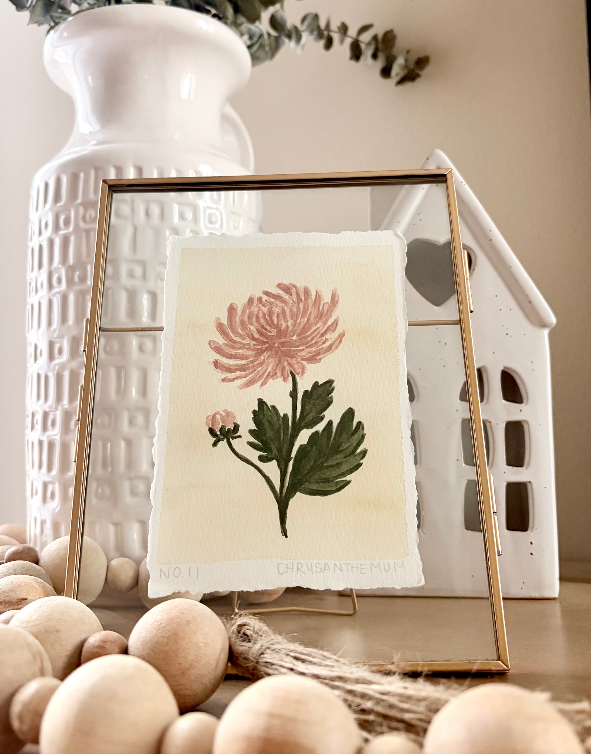 A framed botanical art print featuring a pink chrysanthemum flower and green leaves on a cream textured paper background, styled in a home decor setting with a gold frame, white ceramic vase, and wooden beads.
