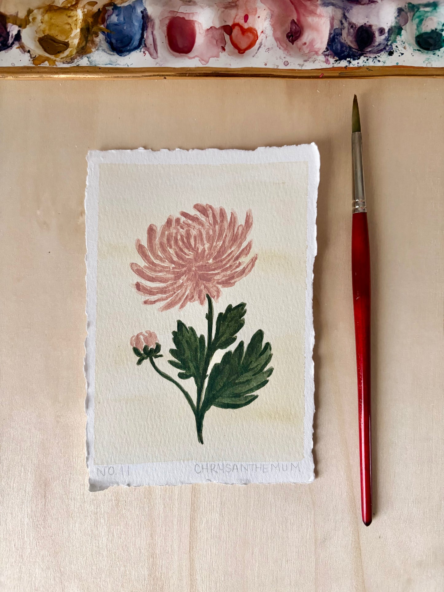A 4x6 watercolor painting of a pink chrysanthemum flower with green leaves on textured paper, placed on a light wood surface next to a red-handled paintbrush. A watercolor palette is visible along the top edge.
