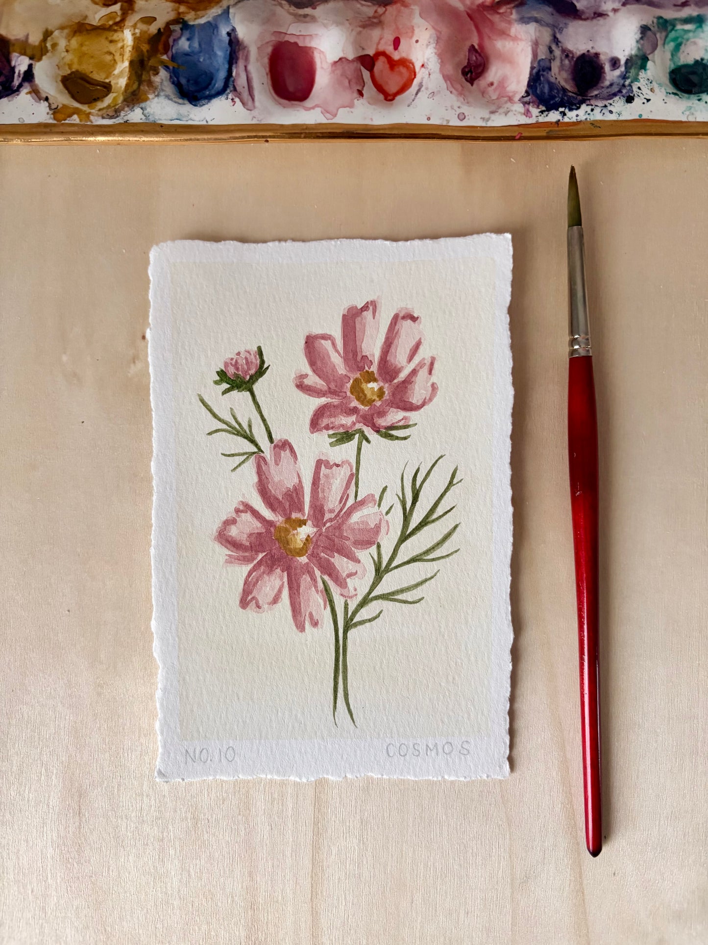A small 4x6 watercolor painting of pink cosmos flowers on textured white paper, displayed on a light wood surface next to a red-handled paintbrush. A painted palette is partially visible at the top.