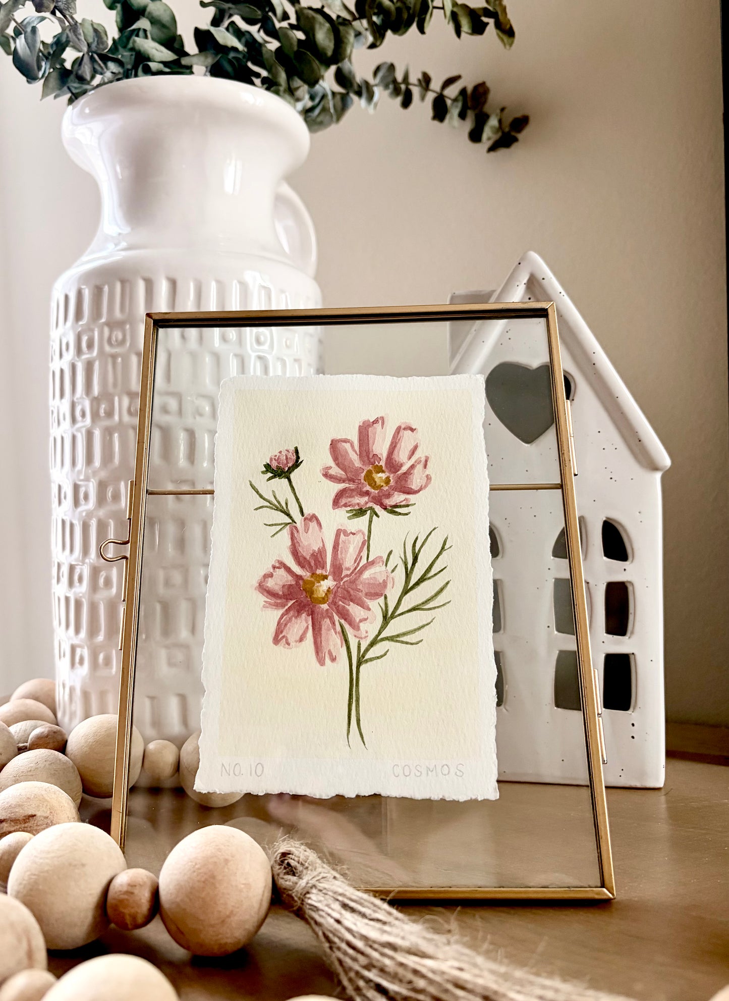 A framed botanical art print featuring pink flowers and green stems on textured cream paper, styled in a home decor setting with a white ceramic vase, wooden beads, and a white house-shaped ceramic object.