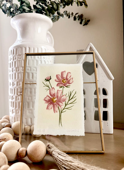 A framed botanical art print featuring pink flowers and green stems on textured cream paper, styled in a home decor setting with a white ceramic vase, wooden beads, and a white house-shaped ceramic object.