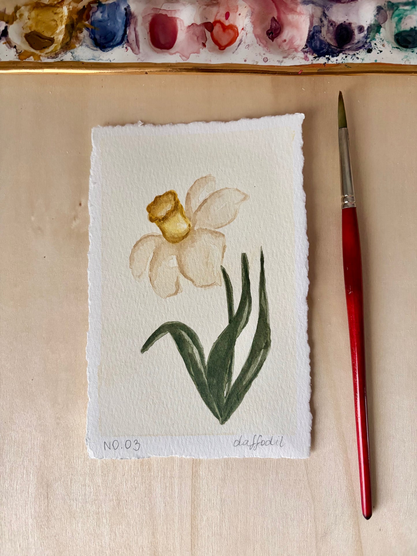 A small watercolor painting of a daffodil on textured white paper, displayed on a light wooden surface next to a red-handled paintbrush. A watercolor palette is partially visible at the top edge.