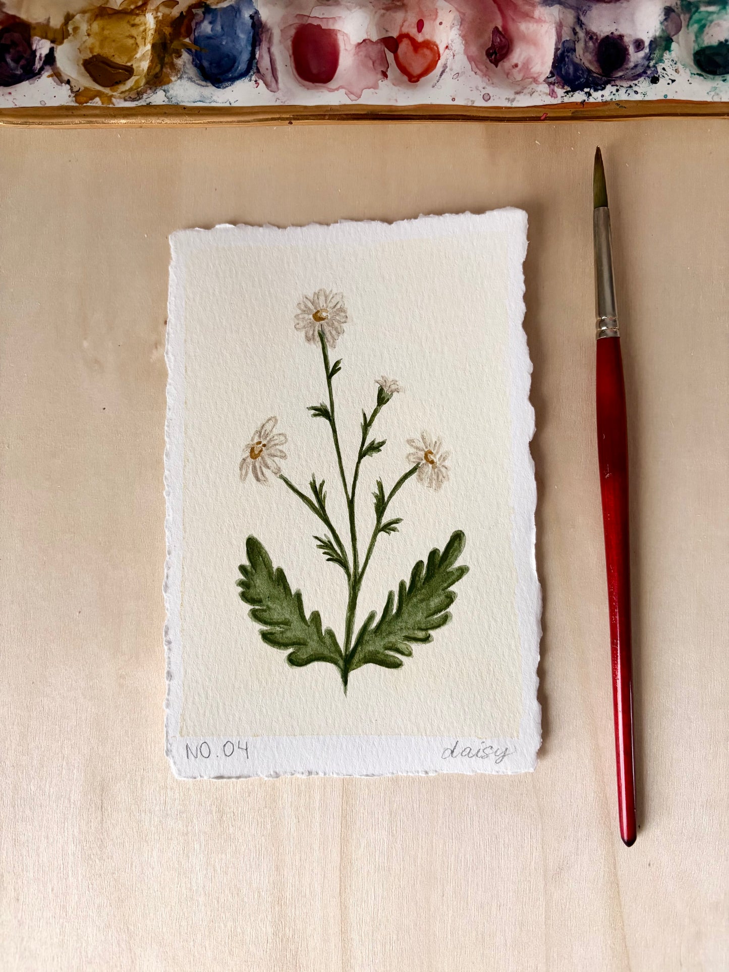 A small botanical art print on textured cream paper showing a green leafy stem with white daisy-like flowers, placed on a light wood surface beside a red-handled paintbrush. A paint palette is partially visible along the top edge.