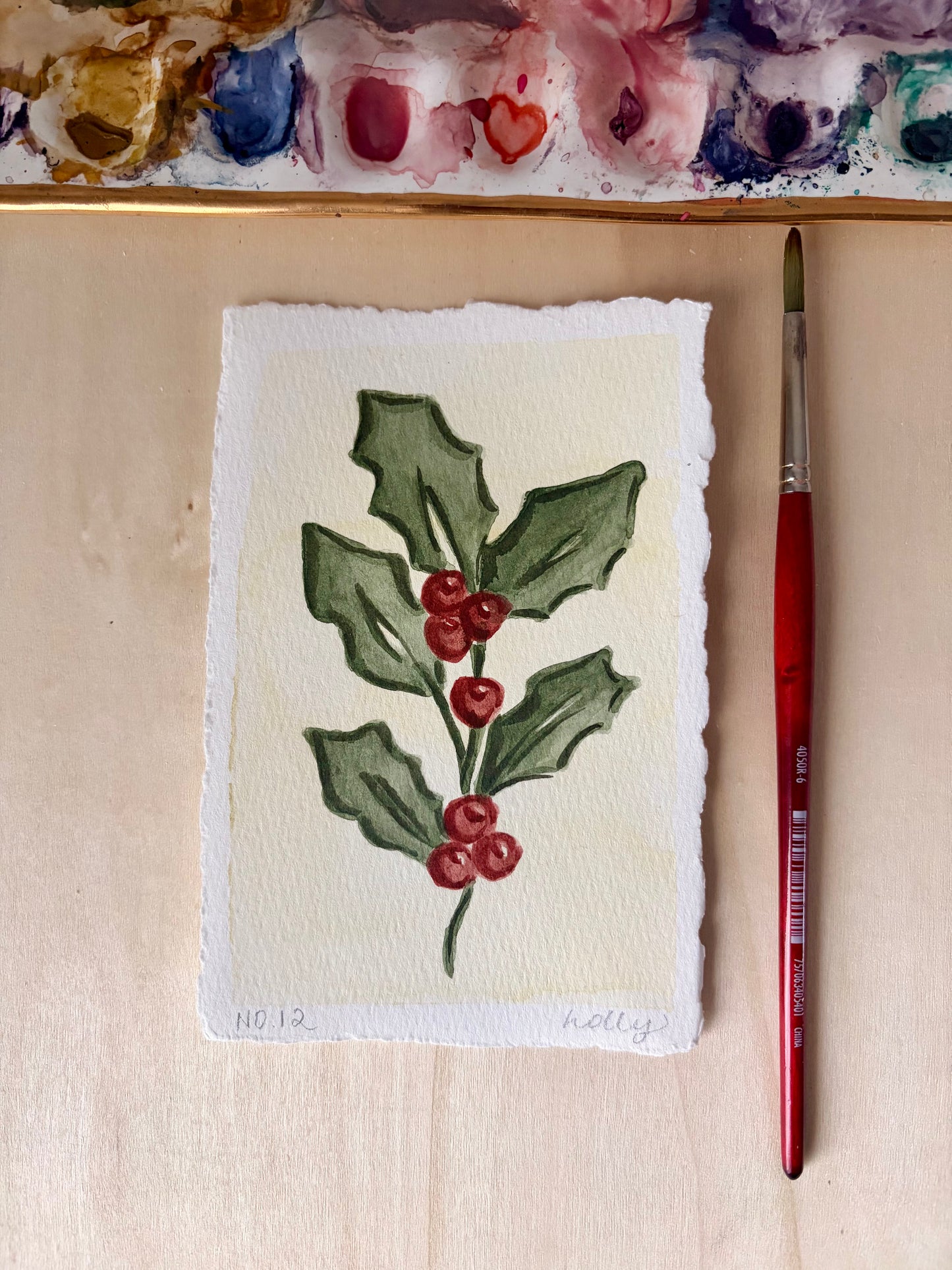 A 4x6 watercolor painting of holly leaves and red berries on textured paper, shown on a light wood surface with a paintbrush and a painted palette visible in the background.