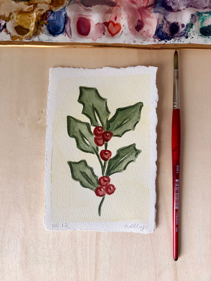 A 4x6 watercolor painting of holly leaves and red berries on textured paper, shown on a light wood surface with a paintbrush and a painted palette visible in the background.