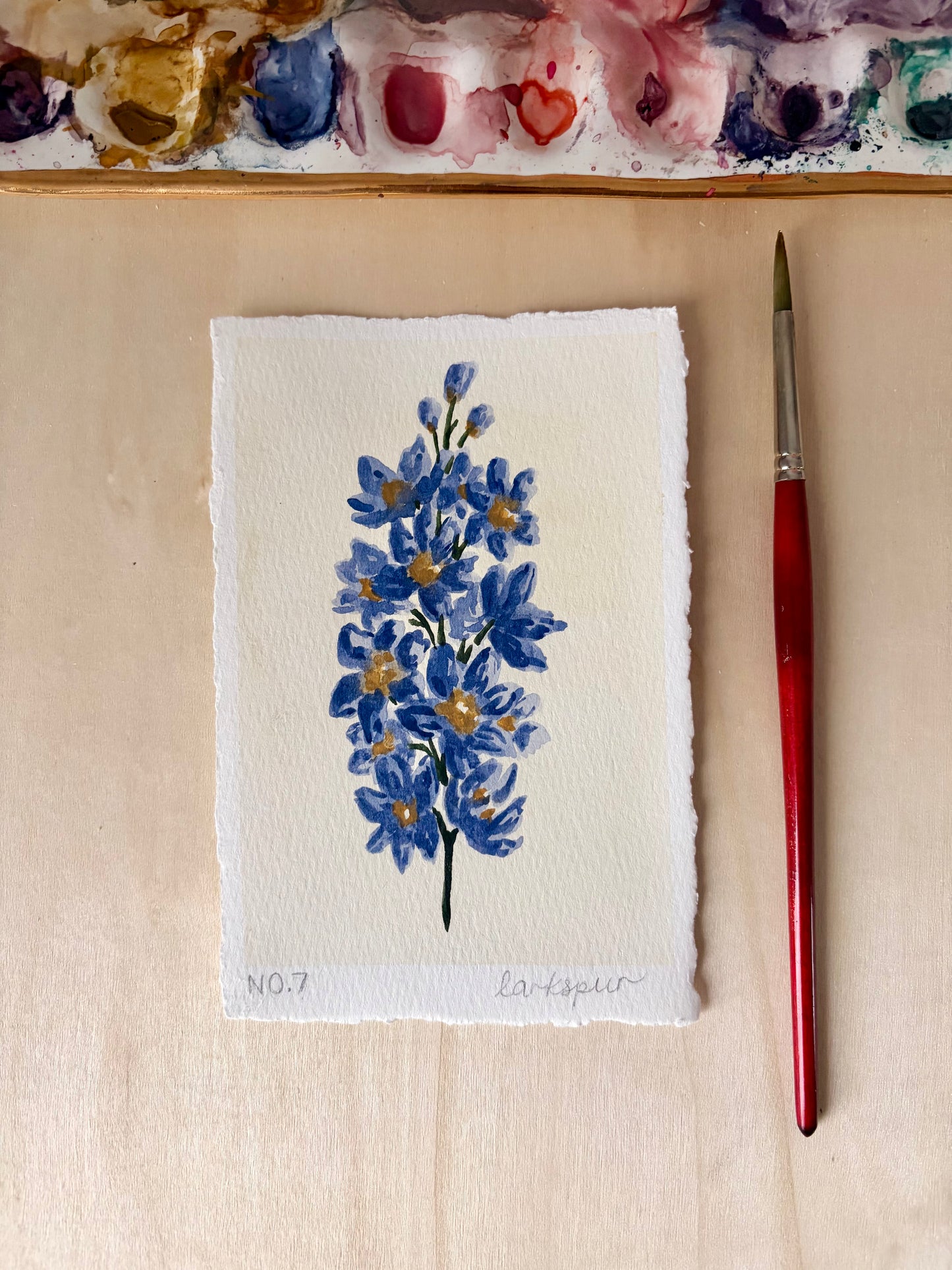 A small square watercolor painting of larkspur blue flowers with yellow centers on textured white paper, placed on a light wood surface next to a red-handled paintbrush. A paint palette is partially visible at the top.