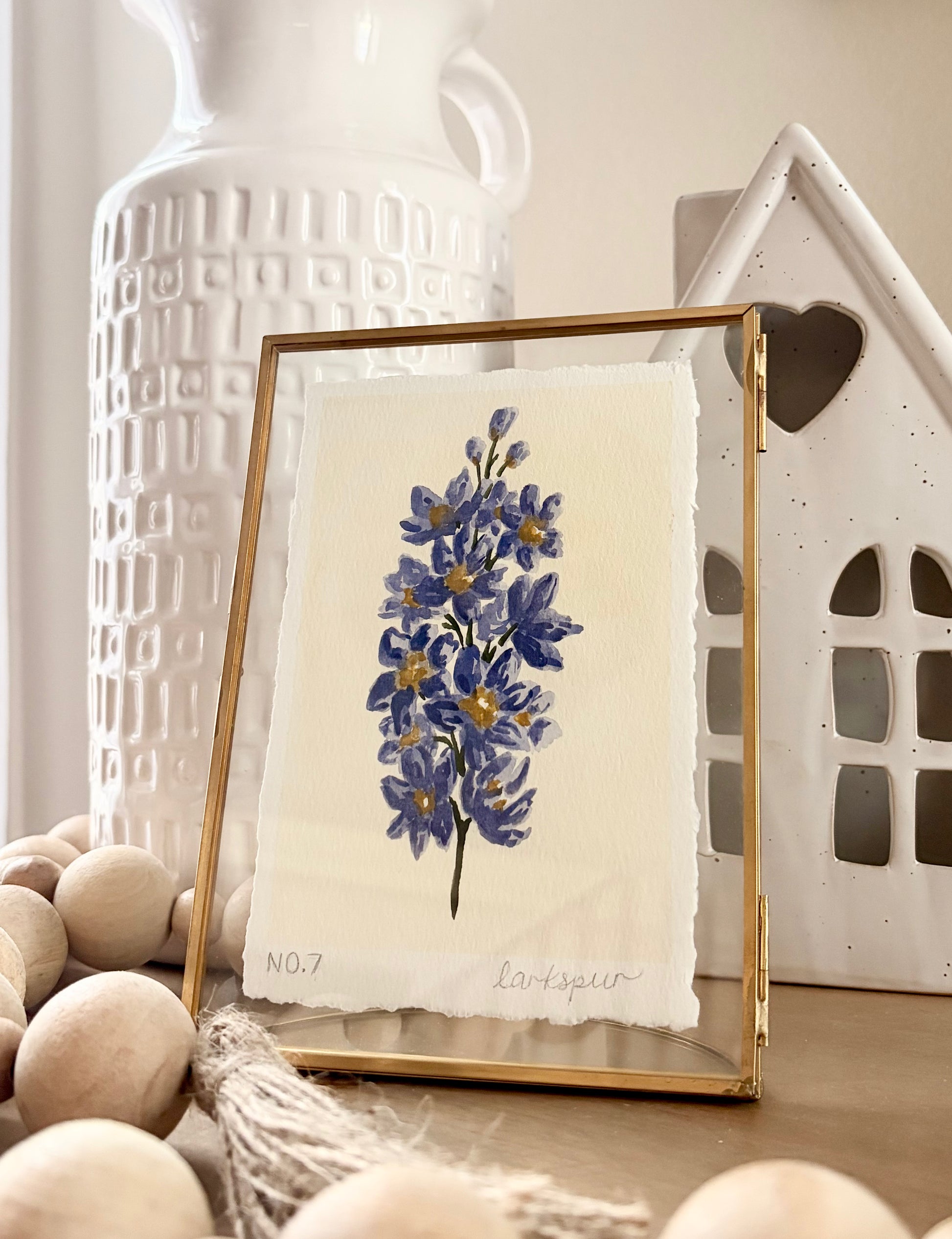 A framed botanical art print featuring blue delphiniums on textured cream paper, set in a gold frame. The frame is styled on a wooden surface with decorative wooden beads, a white textured ceramic vase, and a white ceramic house in the background.