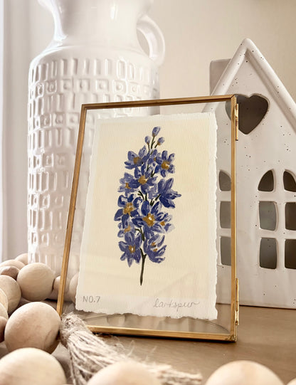 A framed botanical art print featuring blue delphiniums on textured cream paper, set in a gold frame. The frame is styled on a wooden surface with decorative wooden beads, a white textured ceramic vase, and a white ceramic house in the background.