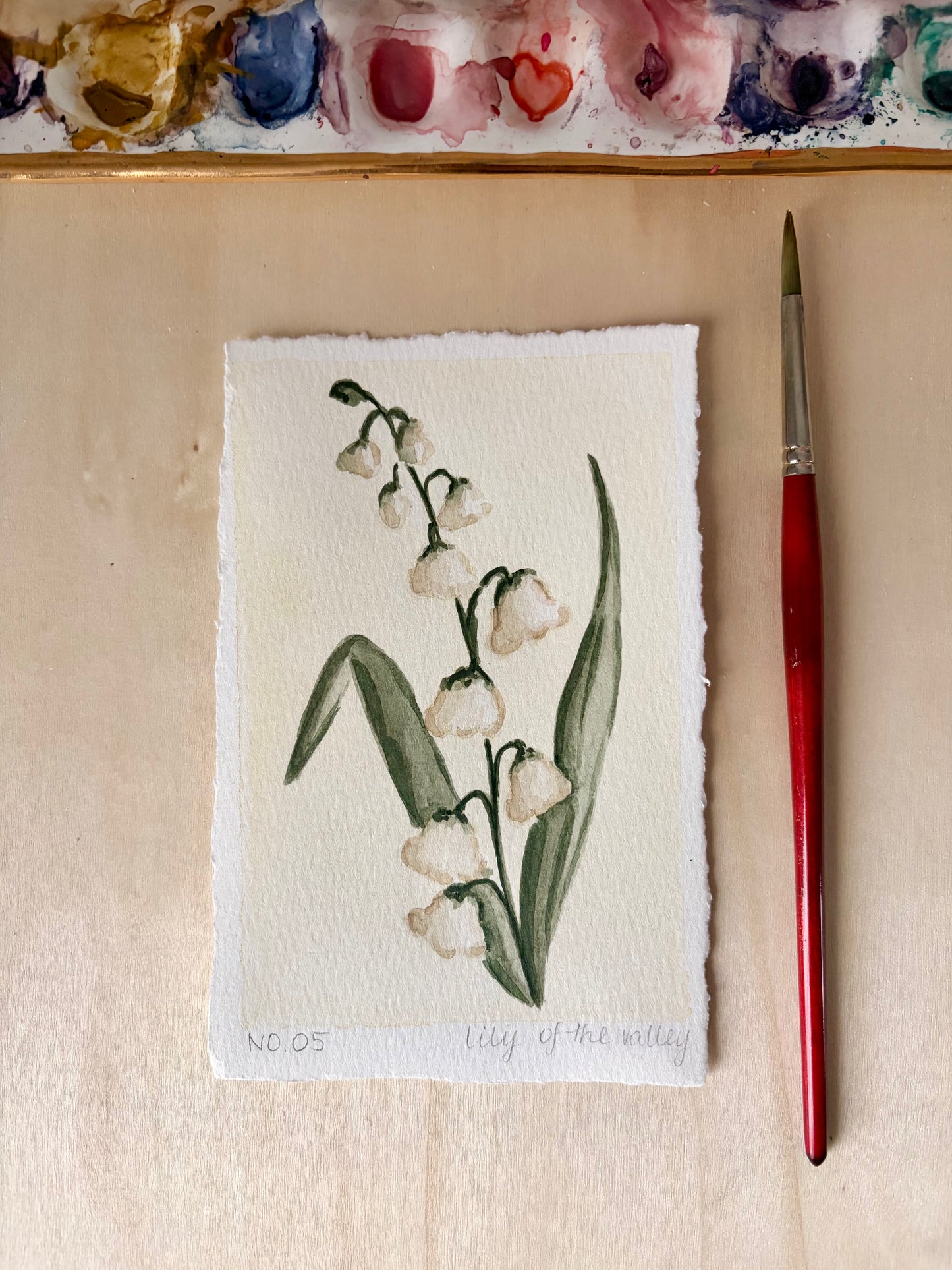 A hand-painted watercolor painting of a lily of the valley on textured white paper, displayed on a light wood surface next to a red-handled paintbrush, with a watercolor palette visible at the top.