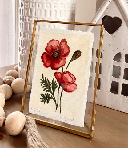 A gold-framed art print featuring a watercolor-style red poppy illustration on textured cream paper, styled on a wooden surface with decorative beads and ceramic objects in the background.