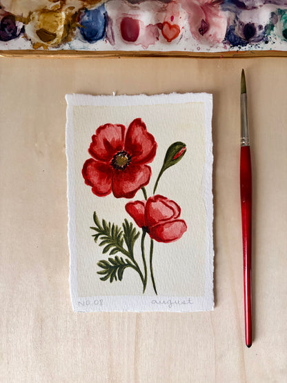 A small watercolor painting of two red poppies with green leaves on textured white paper, displayed on a light wood surface next to a red-handled paintbrush. A paint palette is partially visible at the top.