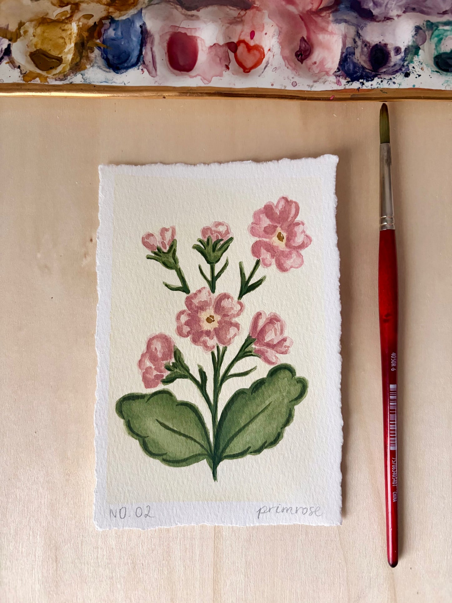 A small watercolor painting of pink primrose flowers and green leaves on textured paper, displayed on a light wood surface with a paintbrush and painted palette visible.