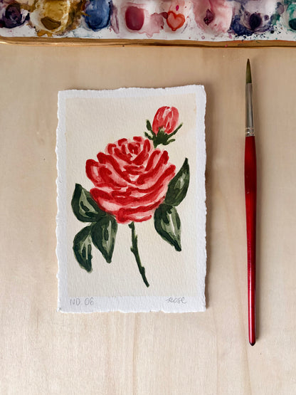A small hand-painted rose artwork on textured white paper, displayed on a light wood surface next to a red-handled paintbrush, with a watercolor palette visible at the top.